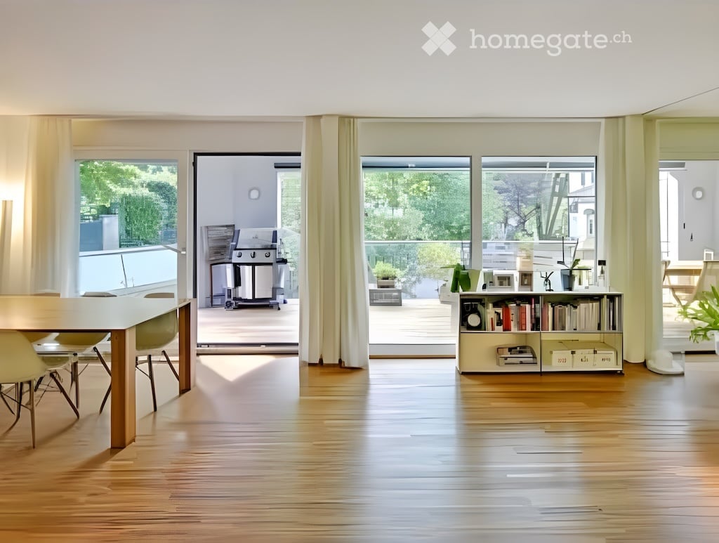 Bright, open-plan living and dining area with wooden flooring, large windows, and a balcony. Includes a dining table with chairs, a bookshelf, and potted plants.