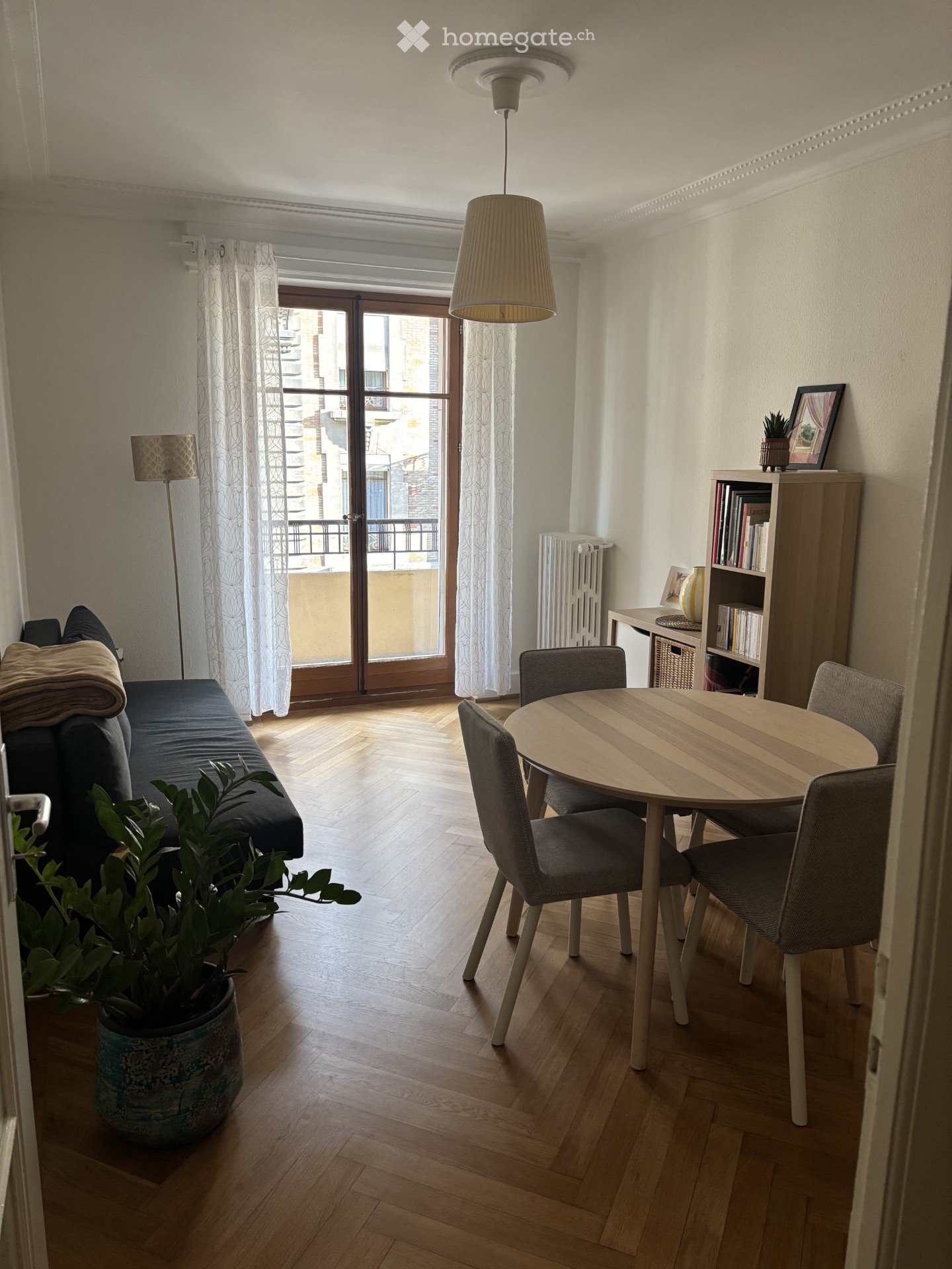 A modern living room with wooden flooring, a sofa, a round dining table with chairs, a bookshelf, a potted plant, and a radiator near the balcony door.