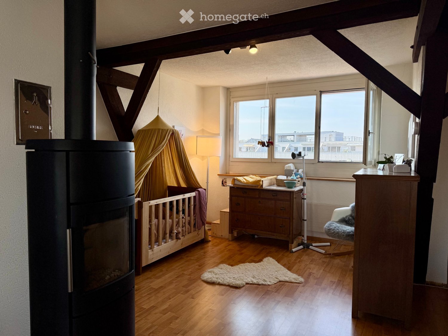 Cozy bedroom with wooden floor, large windows, a cradle, dresser, chair, lamp, and a fireplace.