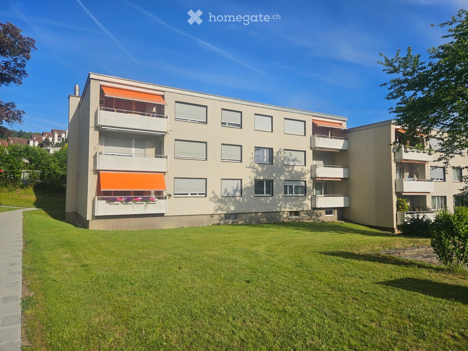 Modern multi-story residential building with balconies, orange awnings, and flower boxes, surrounded by a well-maintained lawn and trees.