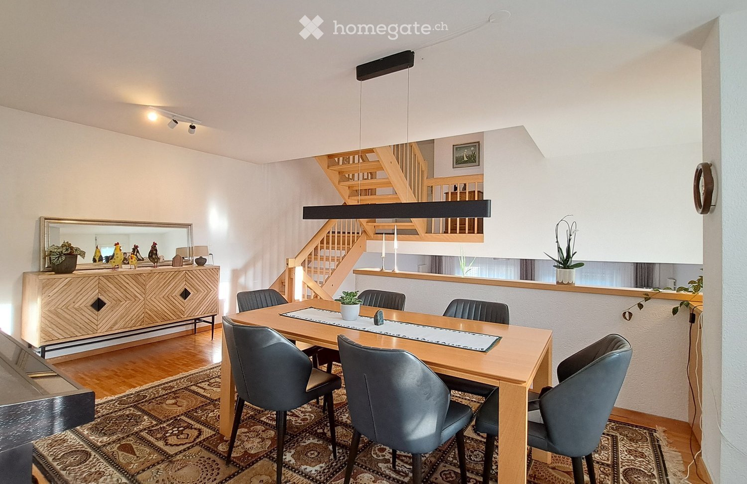 Furnished Room. Dining area with a wooden dining table surrounded by grey chairs, a patterned rug, a wooden cabinet with a mirror, and decorative items including potted plants and candles. A staircase leads to an upper level.