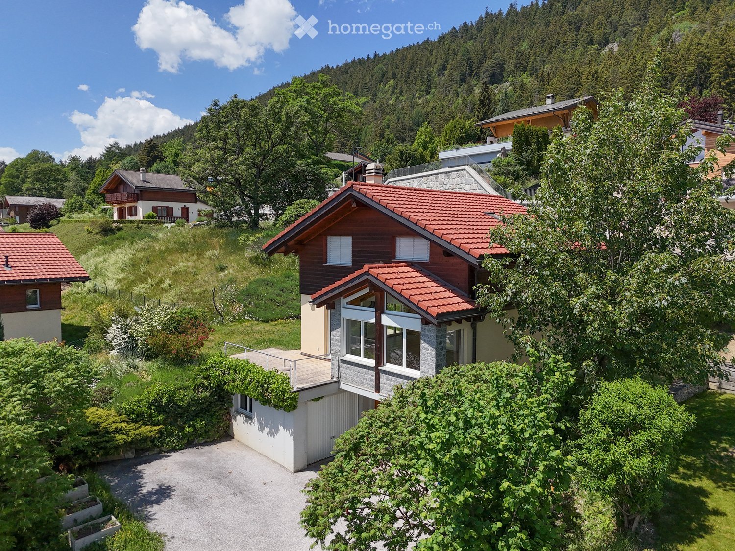 A two-story house with a red-tiled roof, surrounded by greenery and trees in a hilly landscape.