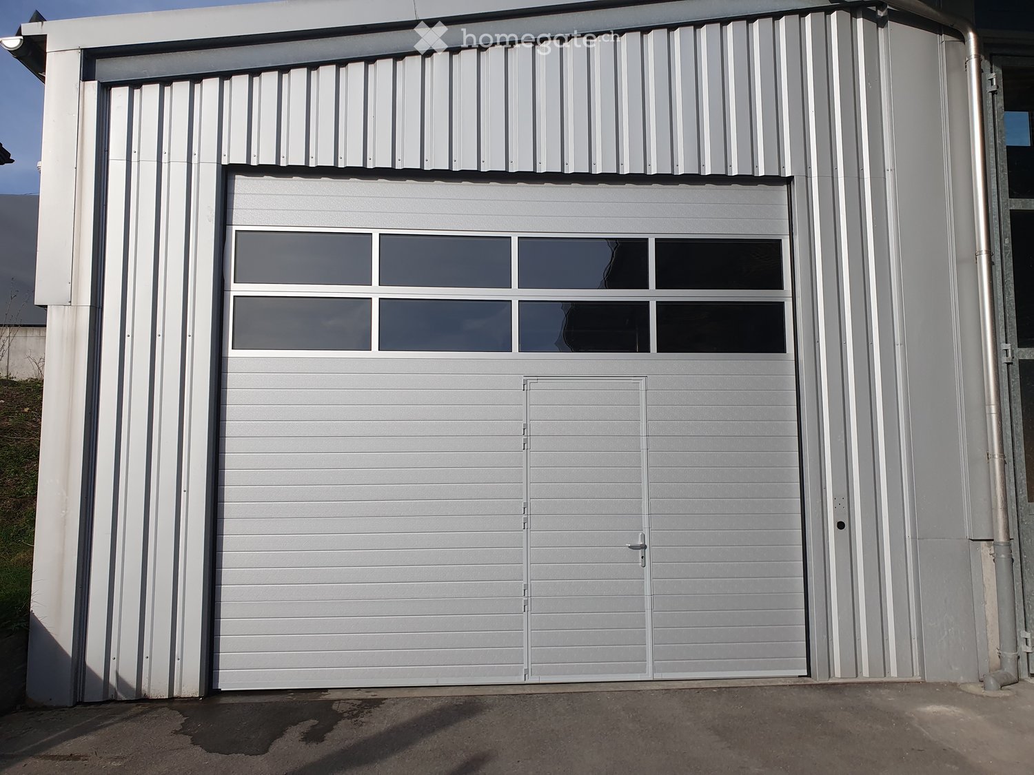 A modern, grey garage with a large, closed garage door featuring a window section at the top. The garage has a sleek, contemporary design with clean lines and a metallic finish.