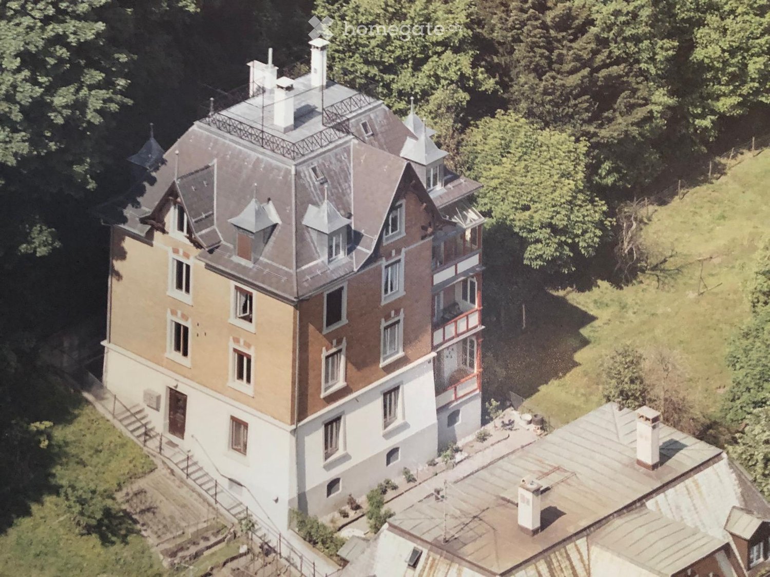 A multi-story residential building with a complex roofline, multiple chimneys, and a mix of beige and red brick facades. The structure is surrounded by greenery and has a raised ground floor with a staircase leading up to the main entrance.
