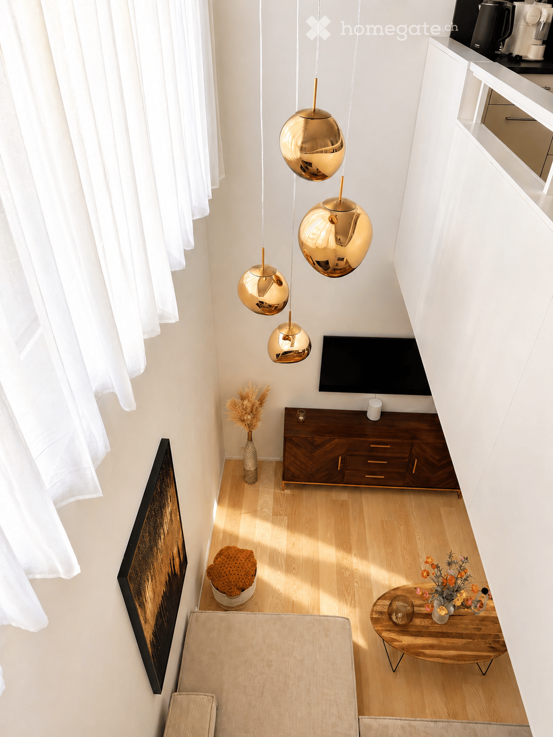 Modern living room with light wooden flooring, white walls, and large windows with white curtains. Features a beige sofa, wooden coffee table with a vase of flowers, TV stand with a flat-screen TV, and decorative items including a woven basket and a vase