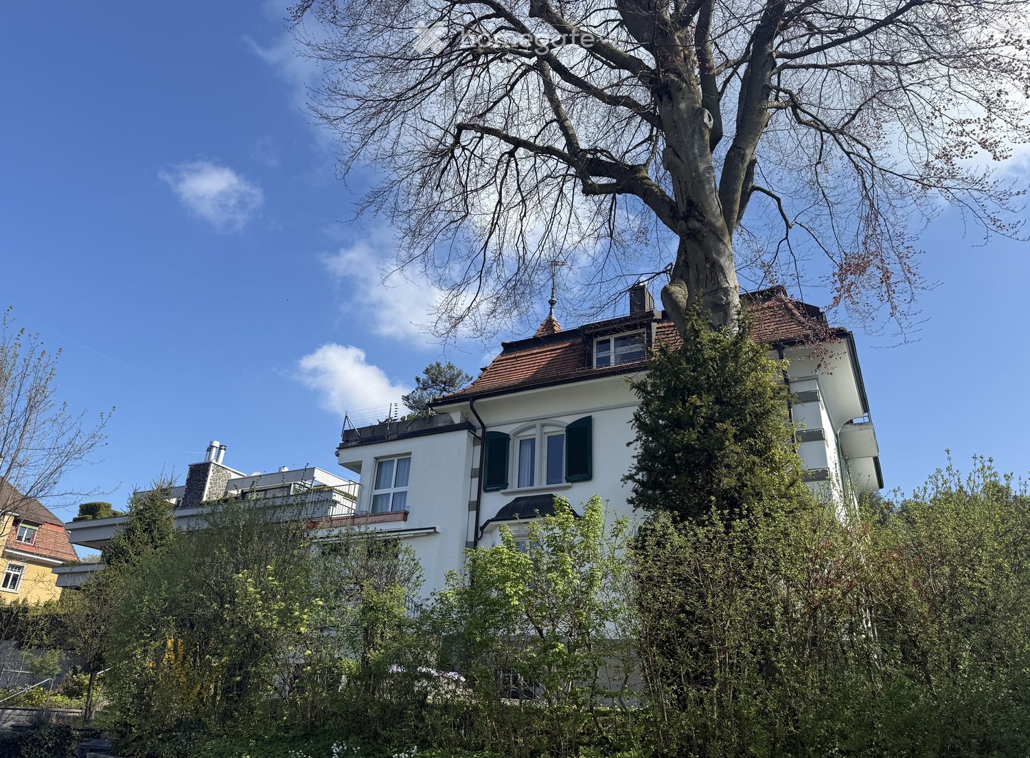 A multi-story white house with a red-tiled roof, surrounded by trees and greenery. The house features multiple windows with shutters and a small balcony on the upper floor.