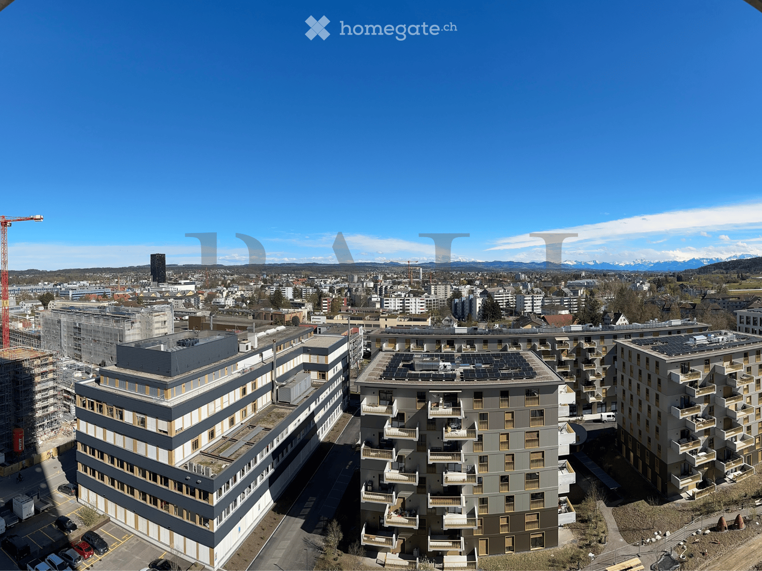 Aerial view of a city with multiple buildings, some under construction, solar panels on rooftops, and a clear view of the mountains in the distance.