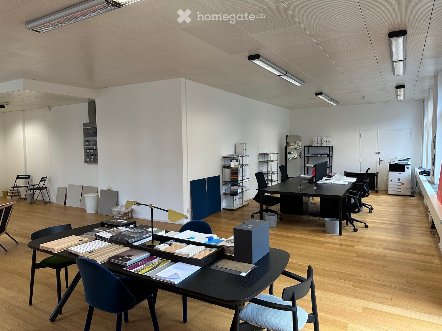 Office space with wooden flooring, multiple desks, chairs, shelves, and office equipment.