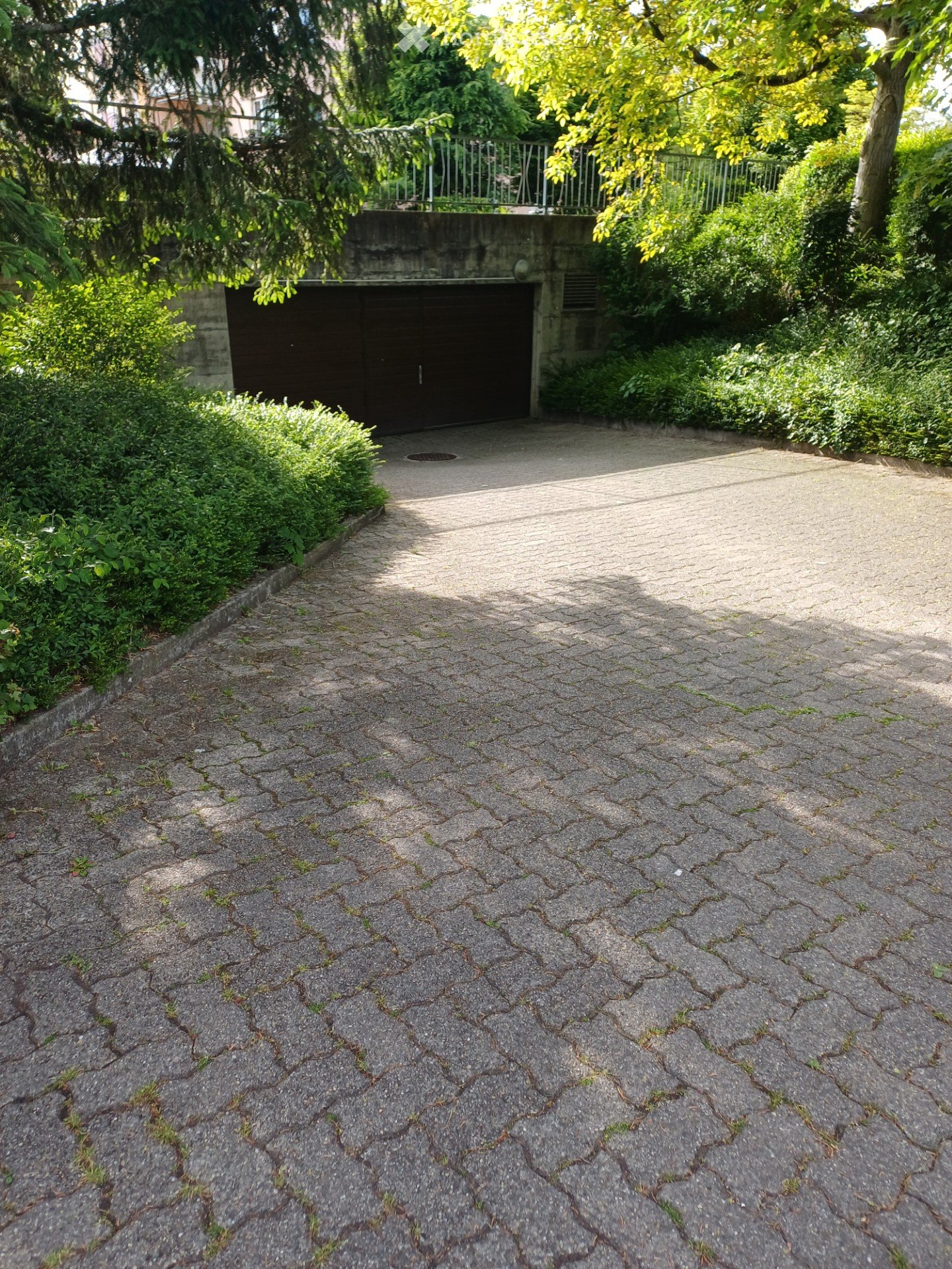 A paved driveway leads to a closed garage, surrounded by greenery and trees, with a concrete wall and metal railing visible in the background.