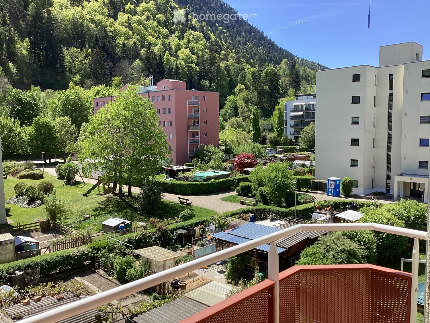 Balcony overlooking a residential area with green spaces, trees, and buildings in the background.