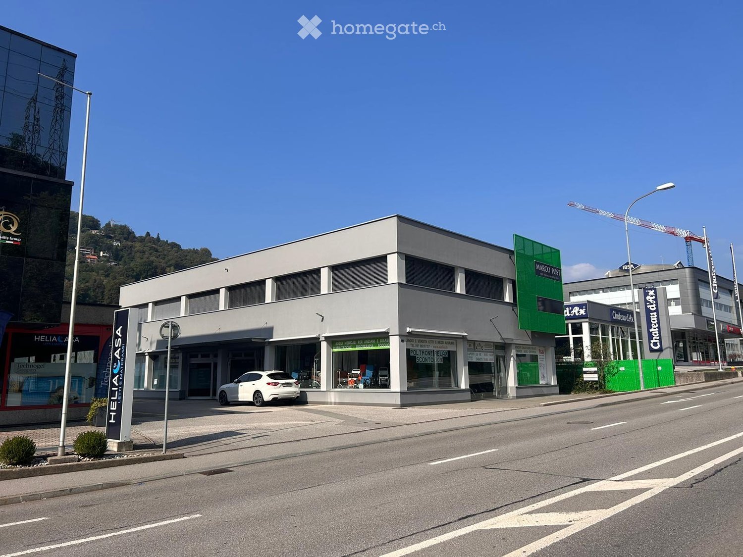 Modern two-story commercial building with signage for 'Marco Post' and 'Heliasa', featuring large windows and a white car parked in front.