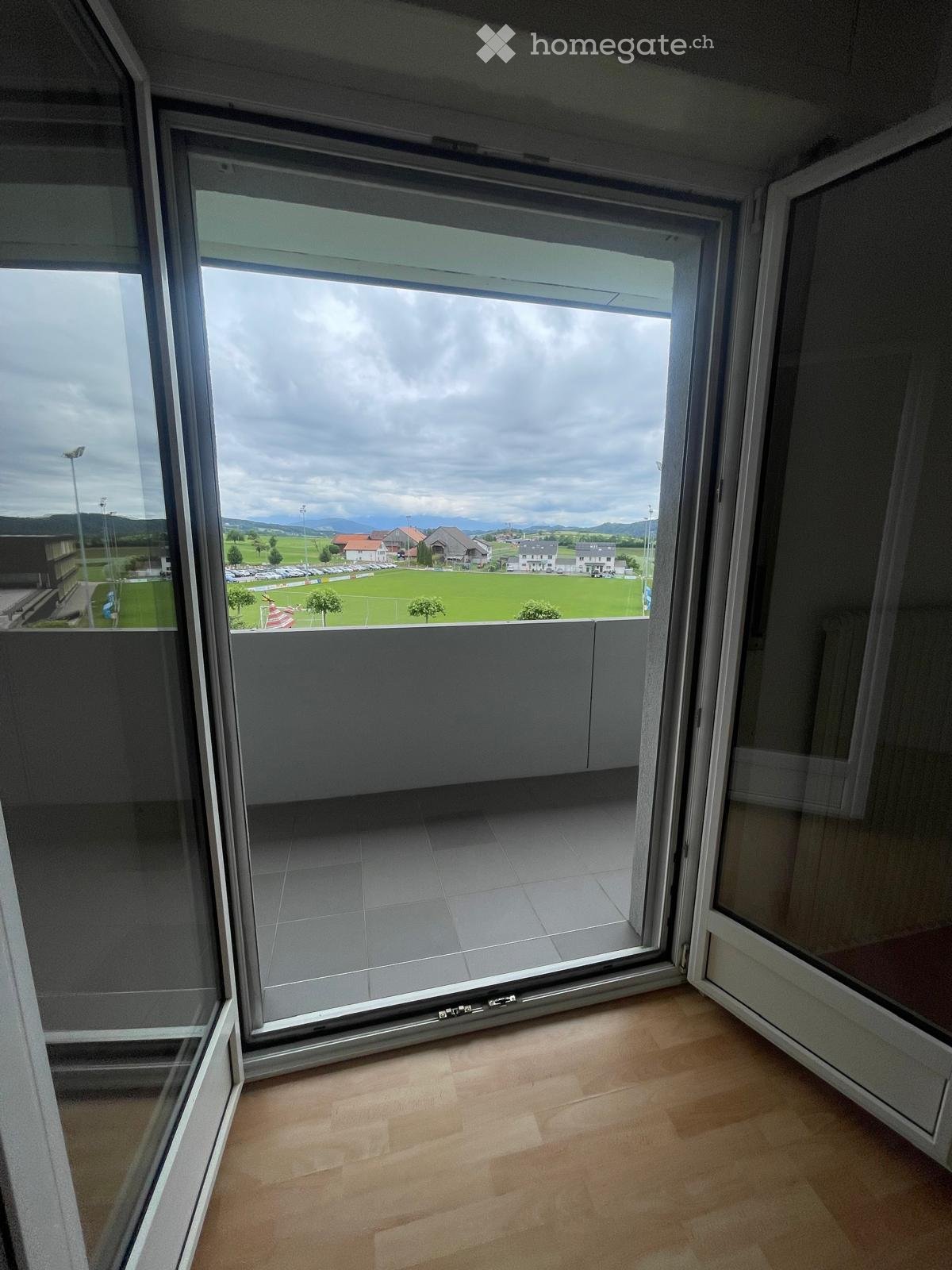 Balcony with large glass doors opening to a view of a green field, distant buildings, and mountains under a cloudy sky.