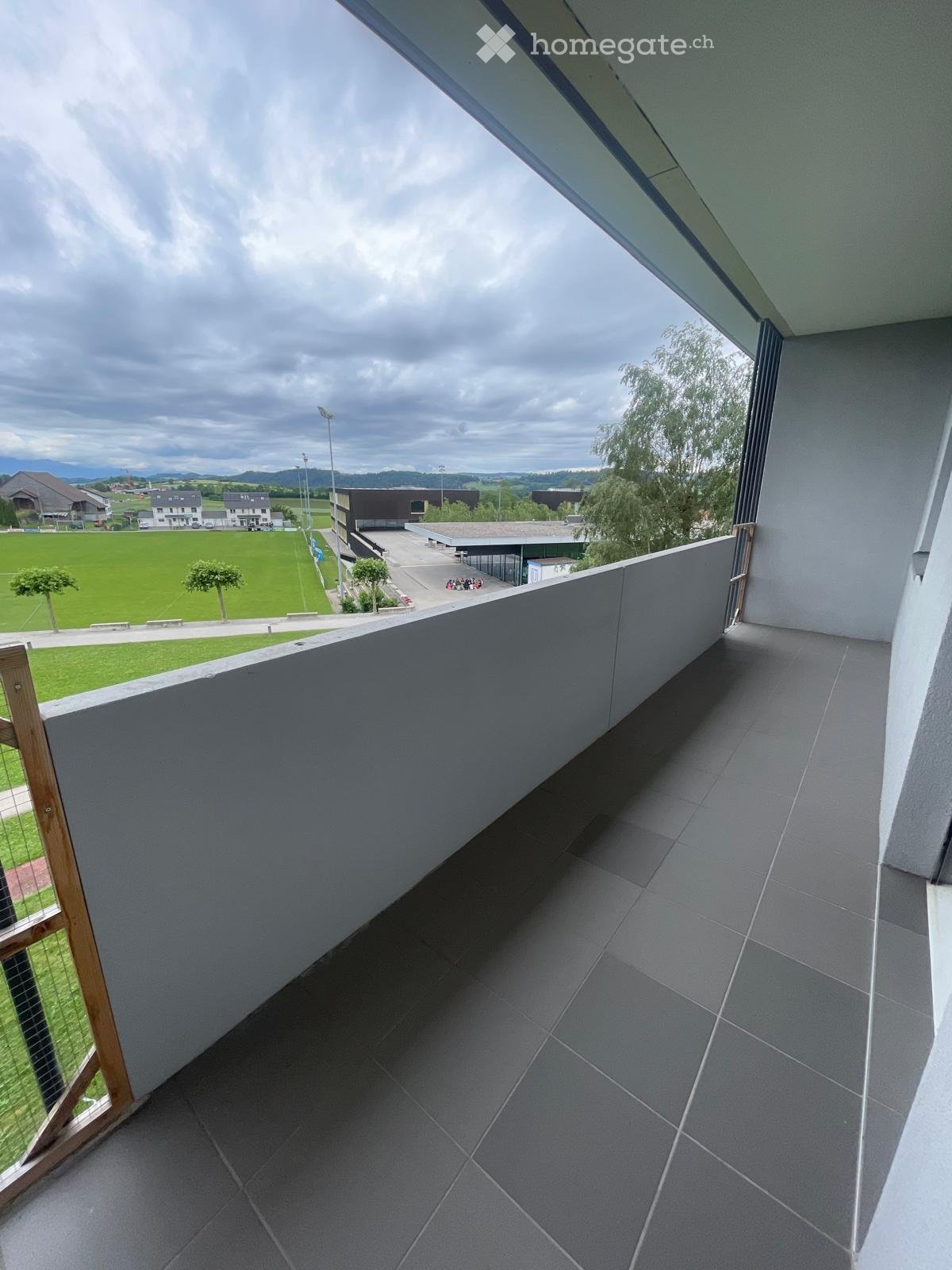 Modern balcony with grey tiled floor, wooden railing, and a view of a green field and buildings.