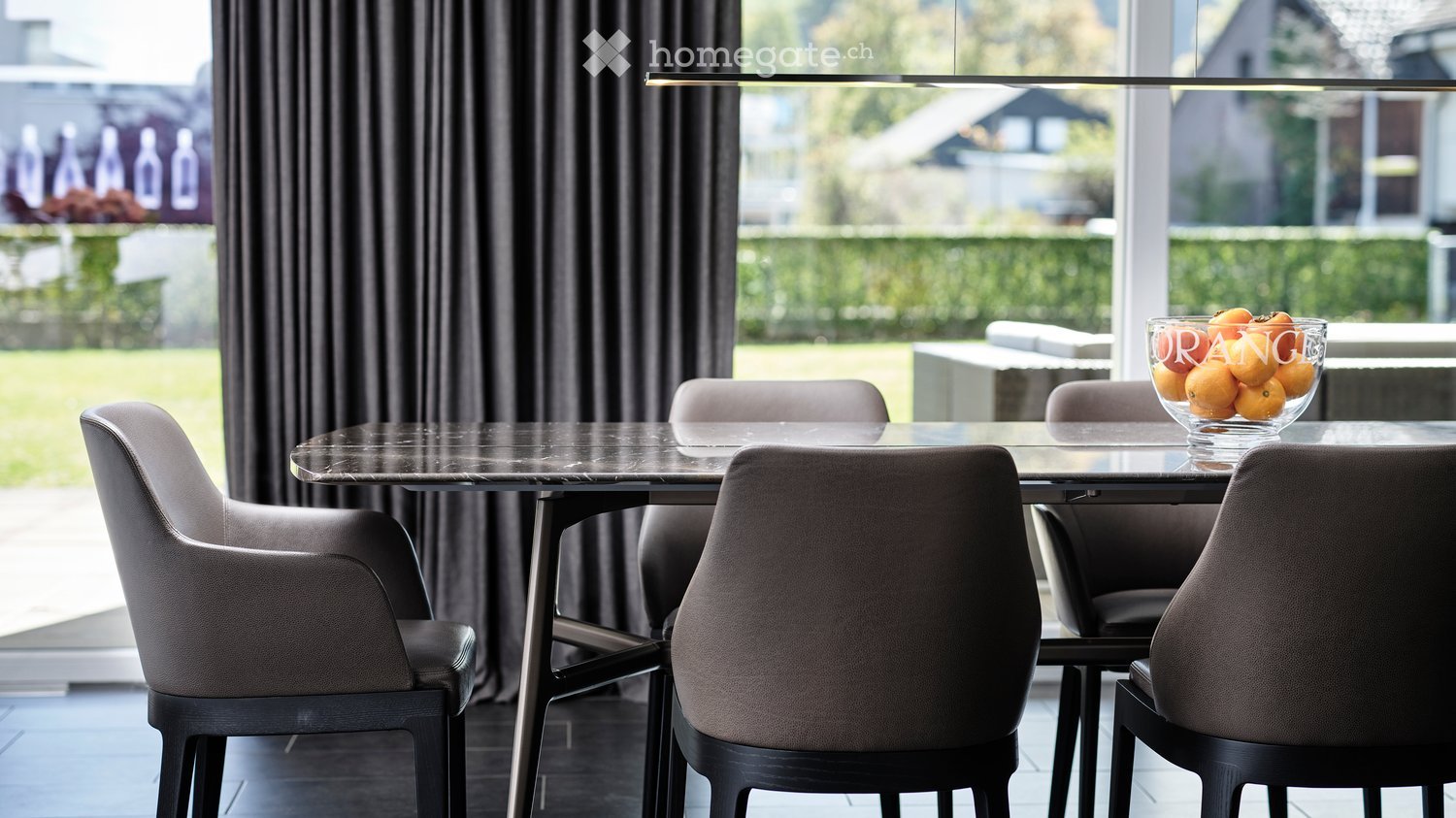 Modern dining room with a sleek glass table, four contemporary chairs, and a bowl of oranges on the table. Large windows with dark curtains offer a view of a green outdoor area.