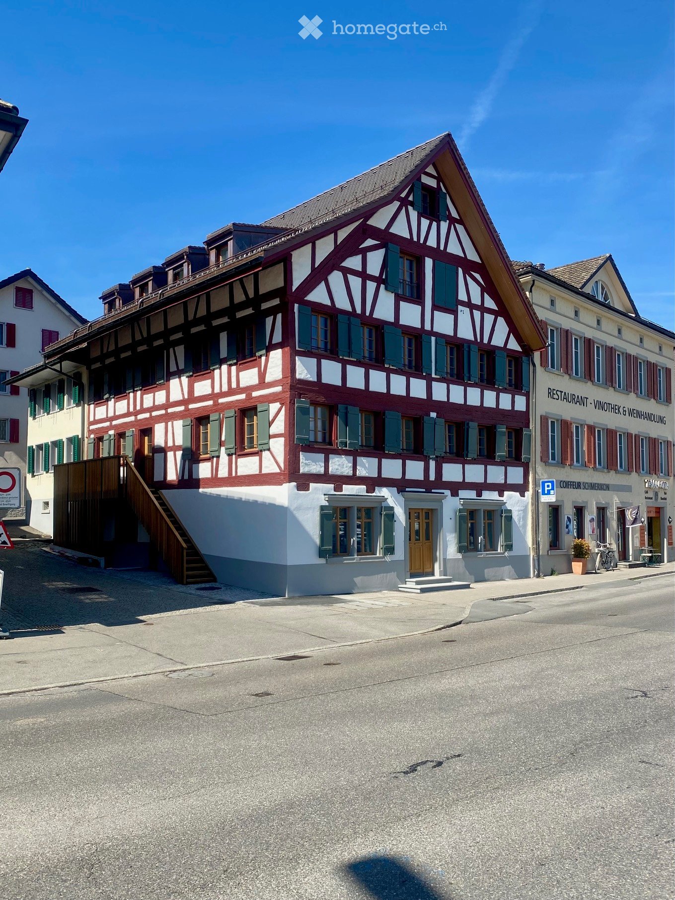 A traditional half-timbered building with red and white facade, green shutters, and a steeply pitched roof, located on a street with adjacent buildings and a parking sign.