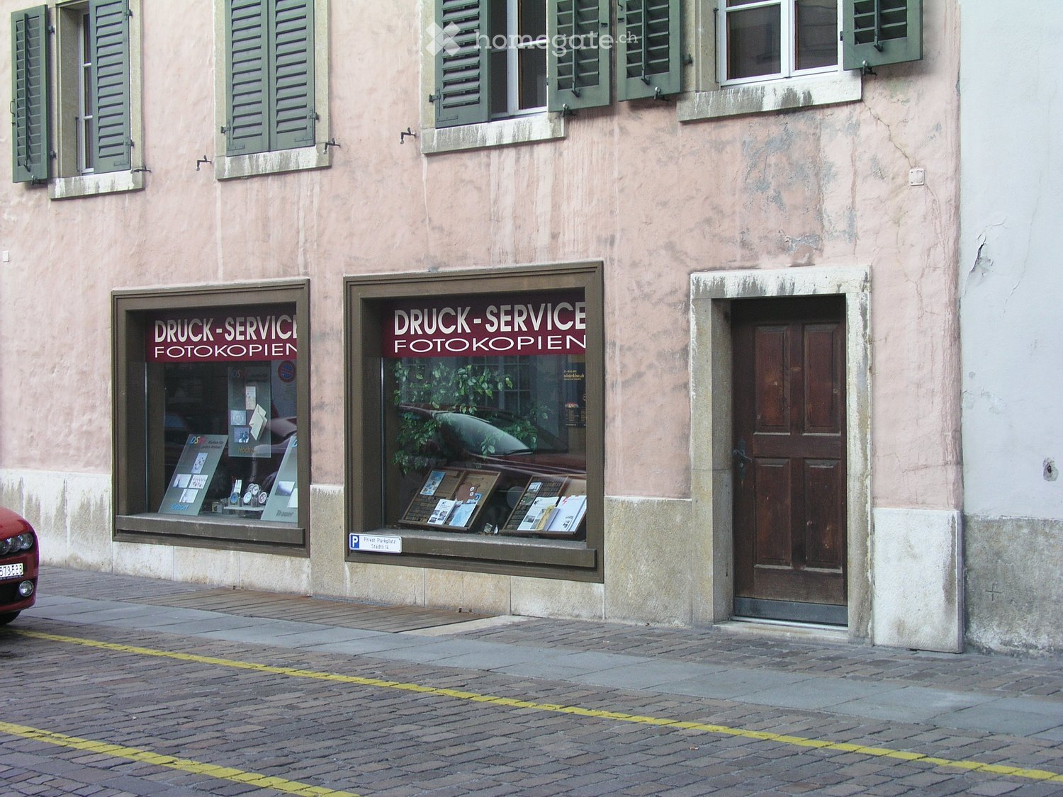 Facade of a two-story building with green shutters, large windows, and a storefront displaying signs for "DRUCK-SERVICE FOTOKOPIEN".