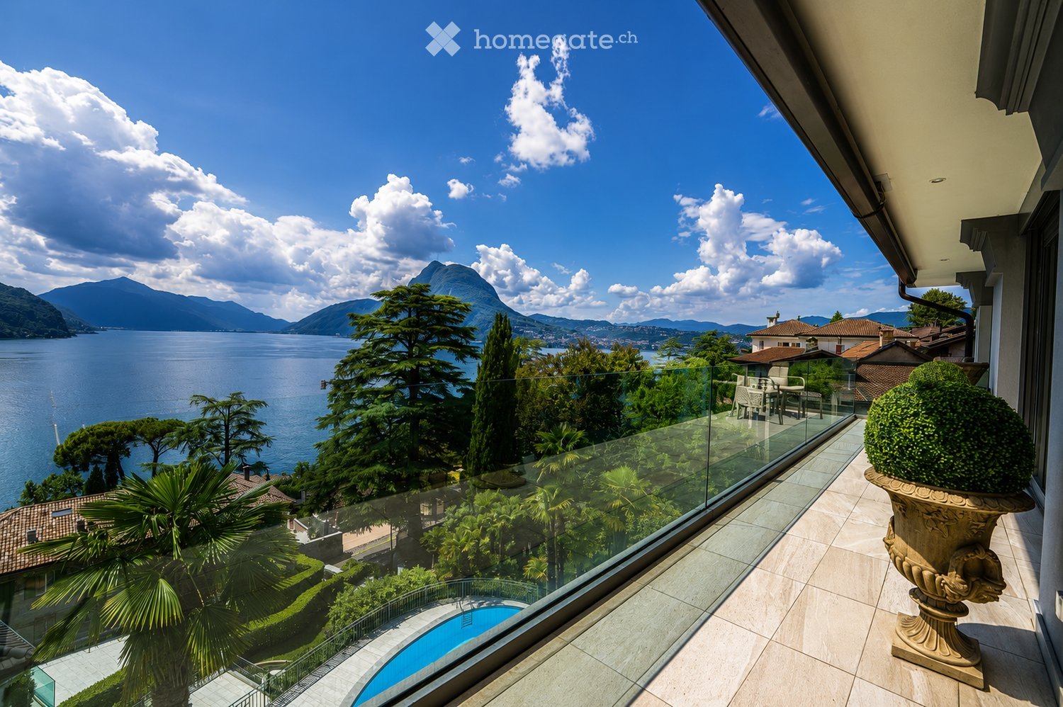 Balcony with a view of a lake and mountains, featuring a glass railing, a small seating area with chairs and a table, and a decorative urn with a plant.