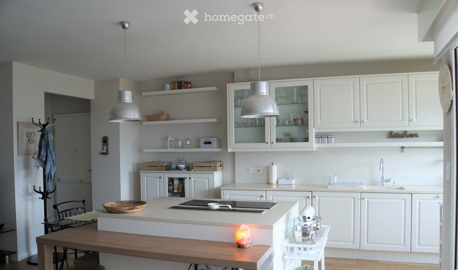 Modern kitchen with white cabinets, glass-fronted upper cabinets, a central island with an induction cooktop, and a wooden dining table with chairs. Shelves hold decorative items and kitchen essentials.