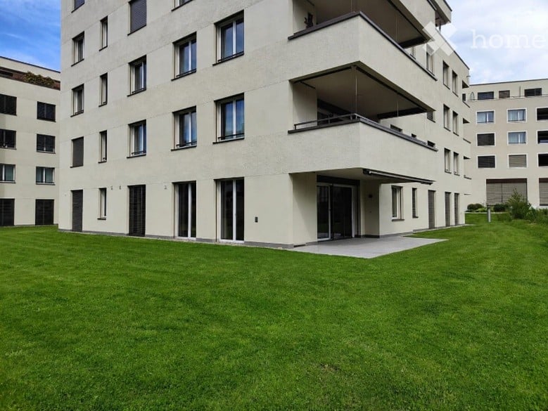 Modern multi-story residential building with balconies and large windows, surrounded by a well-maintained lawn.