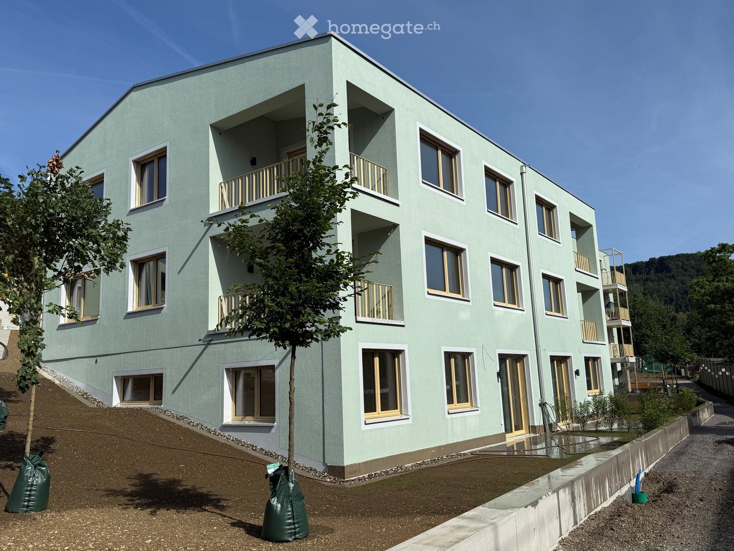 Modern, light green multi-story building with multiple windows and balconies, surrounded by greenery and a raised ground floor.