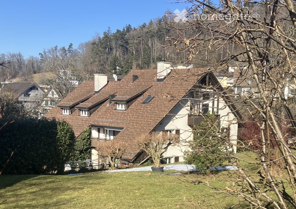 A two-story house with a brown tiled roof, white walls, and multiple windows, situated in a green, hilly area with trees and neighboring houses visible in the background.