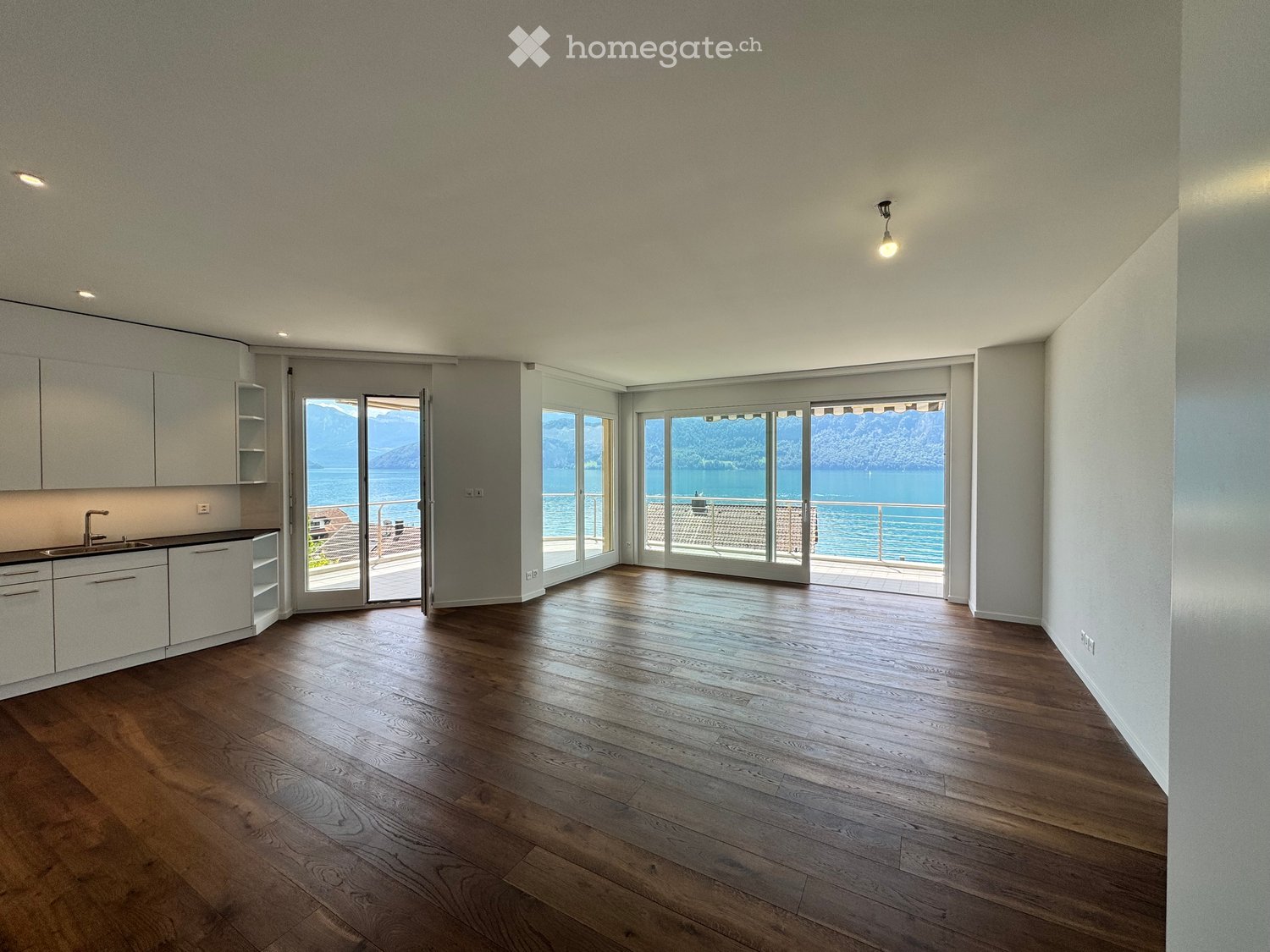 Open-plan unfurnished room with wooden flooring, white walls, large windows, and a view of a lake and mountains. Features a small kitchen area with cabinets and a sink.
