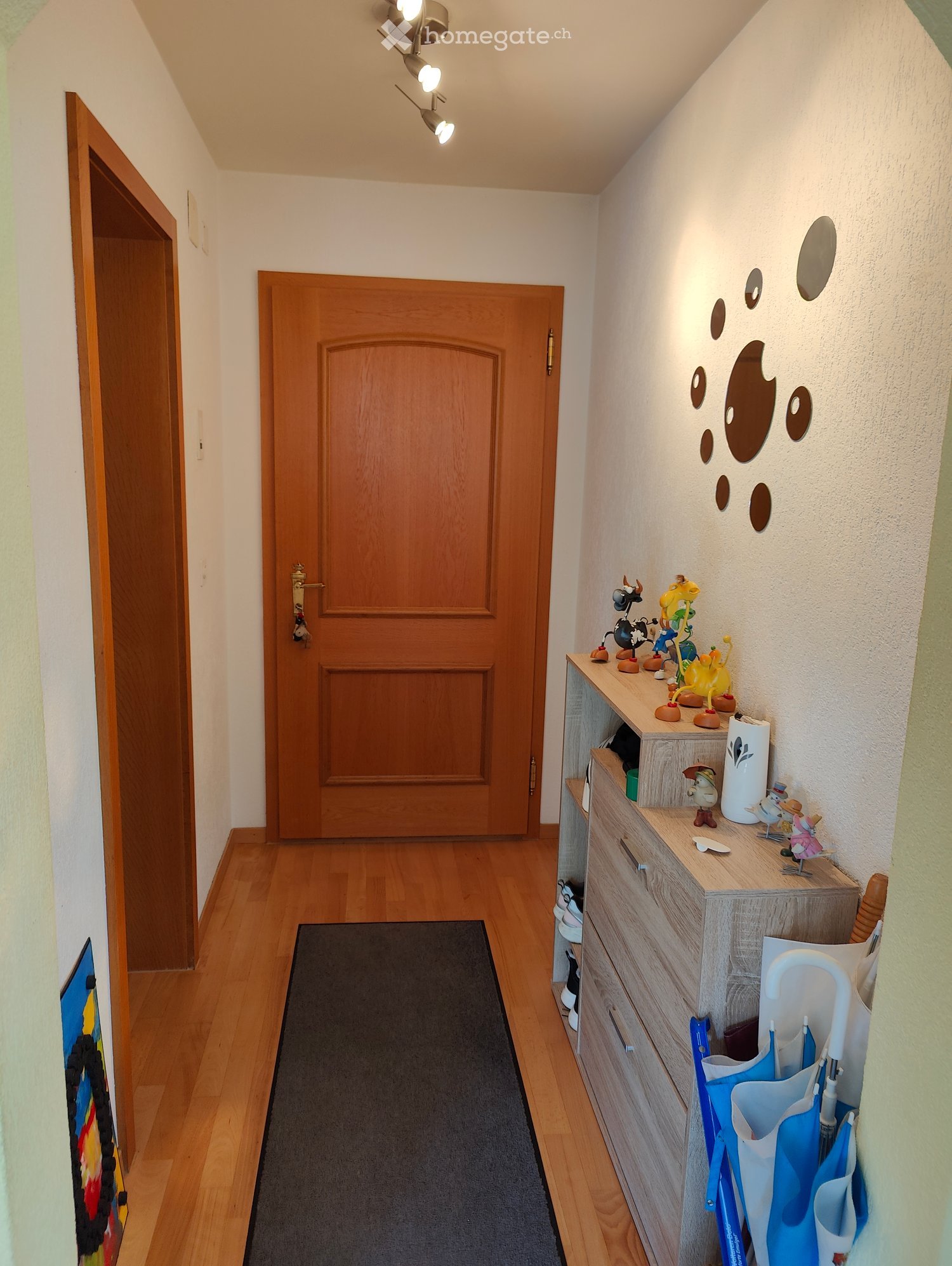 A narrow hallway with wooden flooring, a black runner, and a small wooden cabinet with toys and storage items on top. There are two wooden doors and a wall-mounted light fixture with multiple bulbs.