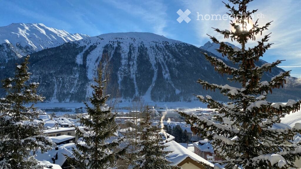 A picturesque winter scene with snow-covered trees in the foreground and a snow-dusted mountain range in the background, with a village nestled in the valley below.