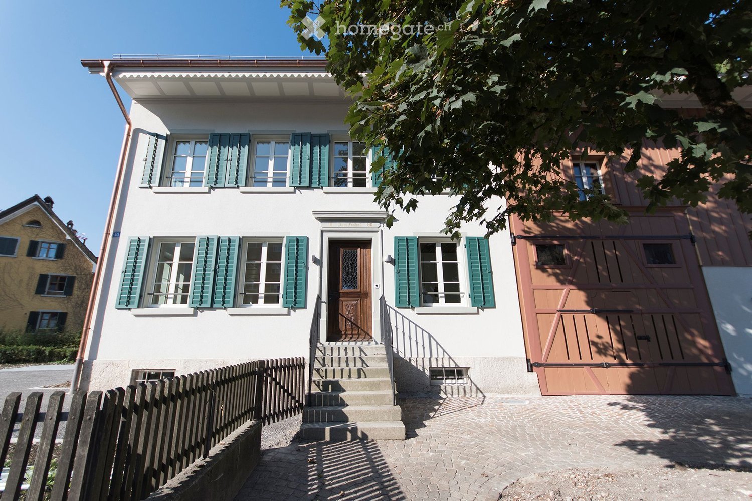 Two-story house with white facade, green shutters, wooden garage door, and steps leading to the entrance.