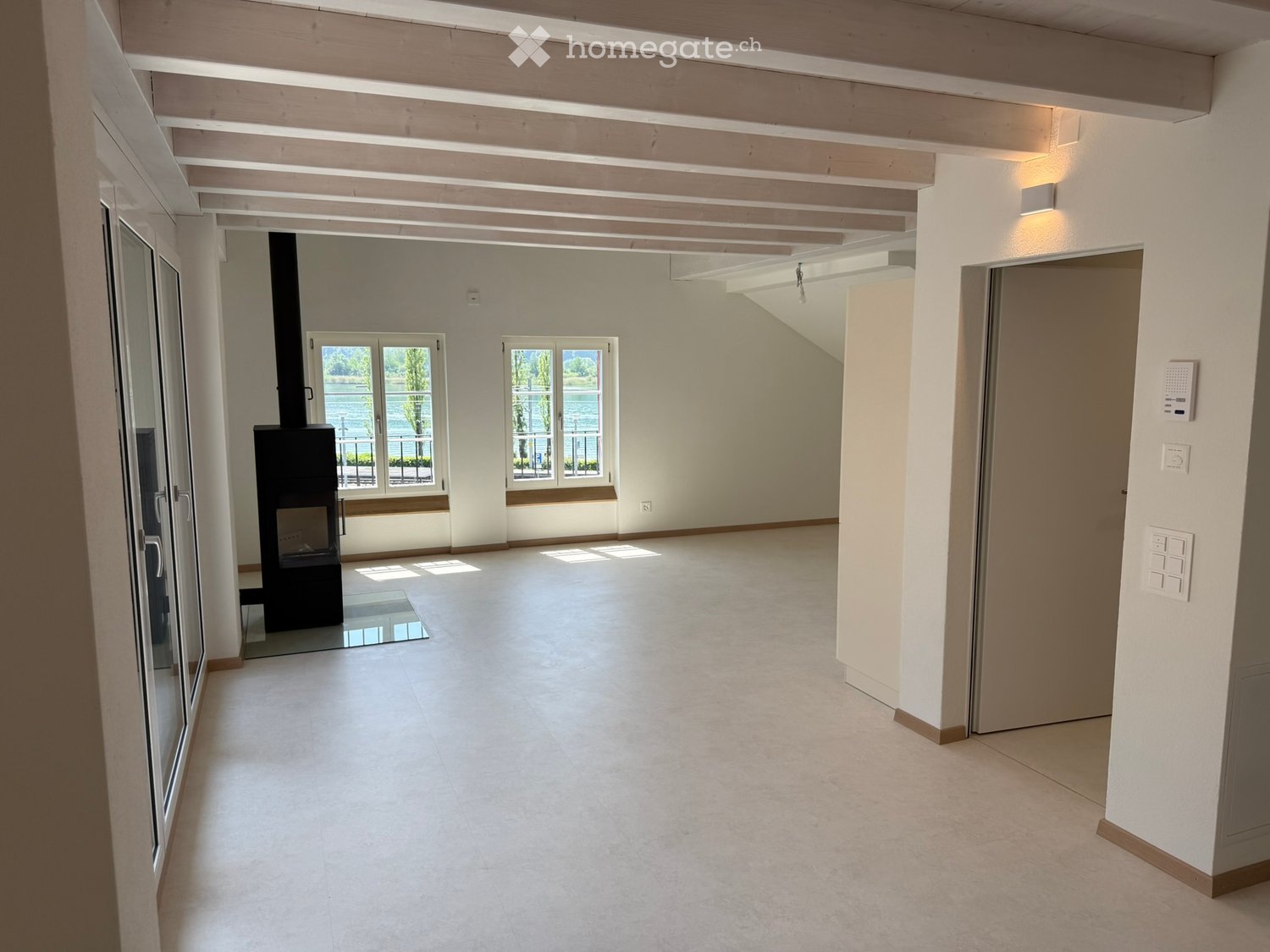 Empty room with white walls, wooden ceiling beams, large windows, and a fireplace. The floor is tiled, and there are no furnishings.