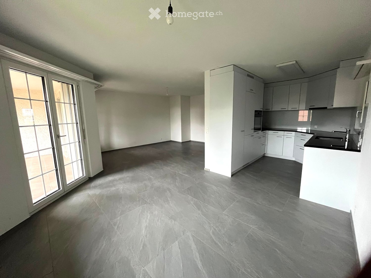 Empty room with grey tiled floor, white walls, large sliding glass door, and a small kitchen area with white cabinets and a countertop.