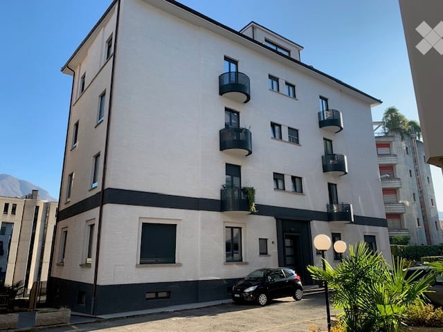 Modern multi-story apartment building with white facade, black trim, and several balconies. A small black car is parked in front.