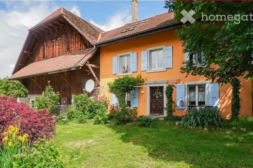 A two-story house with a wooden extension, orange facade, and multiple windows with shutters. The house has a well-maintained garden with various plants and flowers.