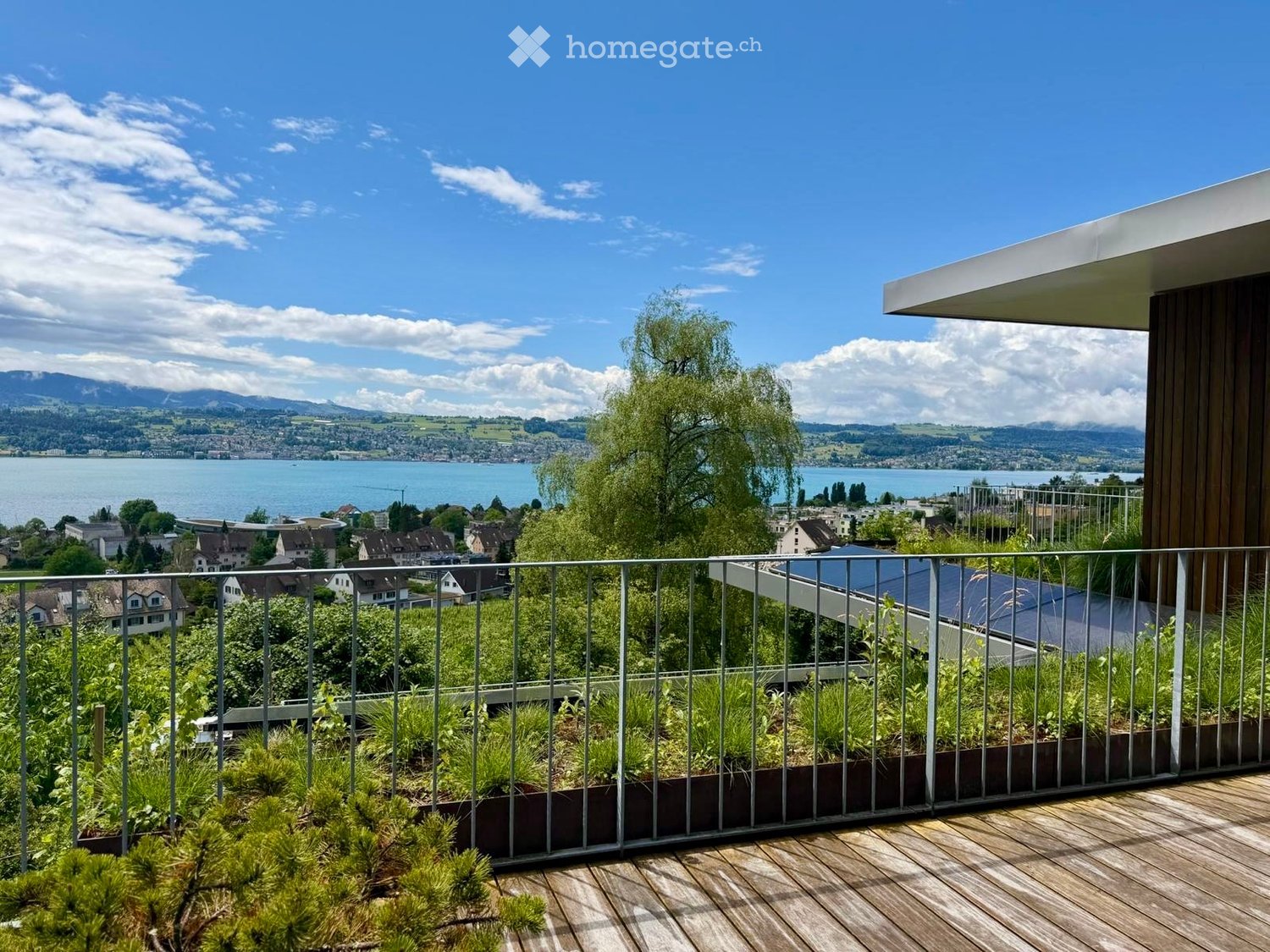 Balcony with wooden decking, metal railing, and lush greenery, overlooking a scenic lake and distant mountains.