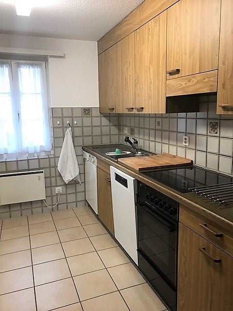 Modern kitchen with light wooden cabinets, tiled backsplash, and stainless steel appliances including a sink, oven, and dishwasher. A window with white curtains provides natural light.