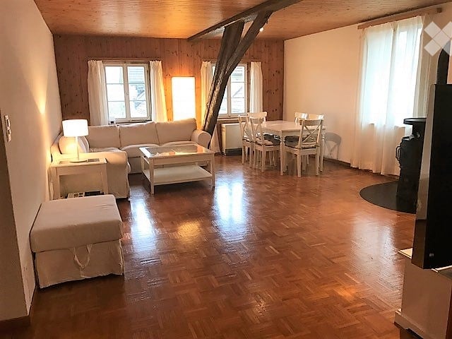 Bright, open living and dining area with wooden floors, large windows, and exposed wooden beams. Features a white sectional sofa, coffee table, ottoman, side table with lamp, and a dining area with a table and chairs.