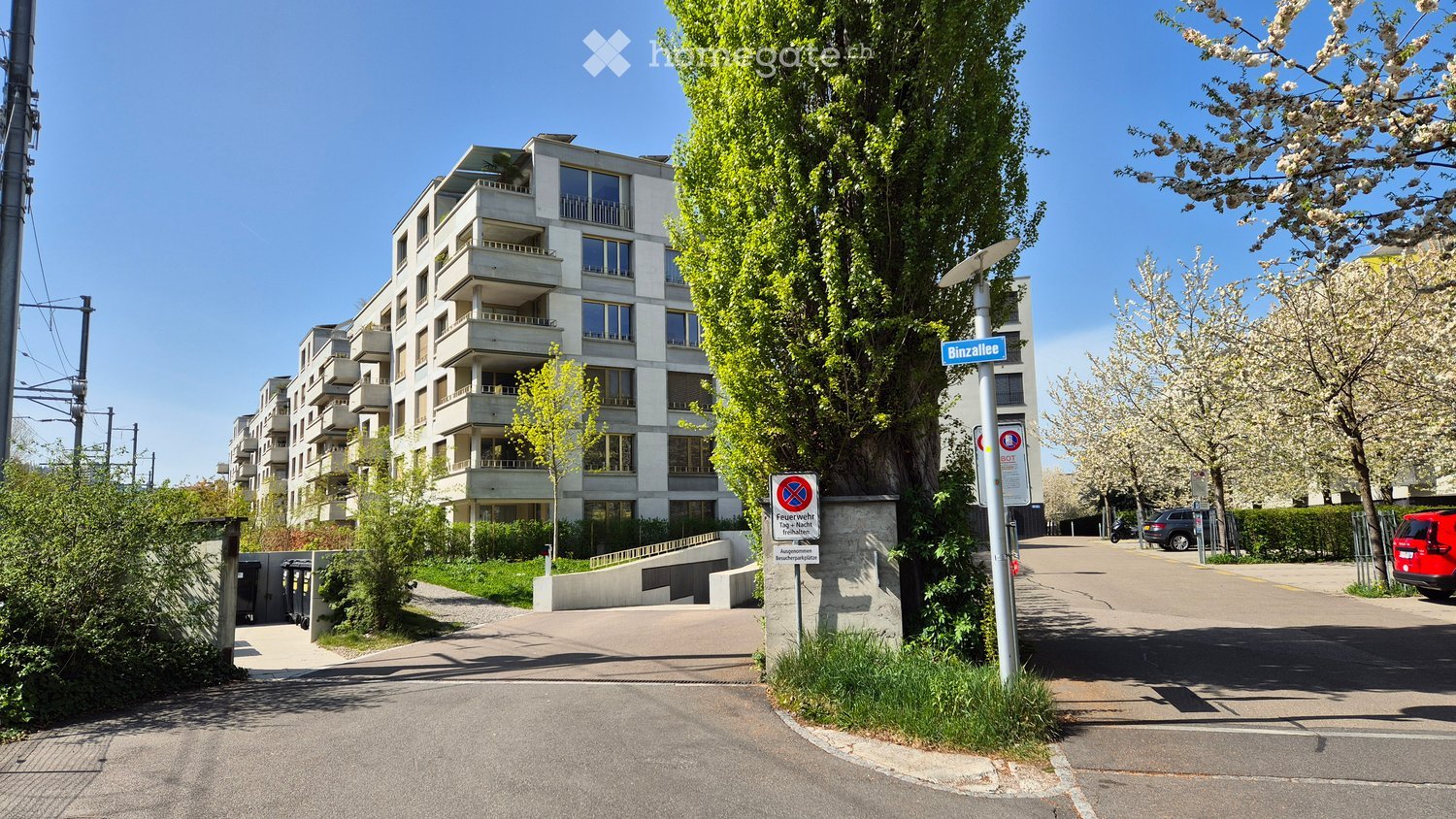 A multi-story residential building with balconies, located on a street with a street sign, trees, and a parking area.