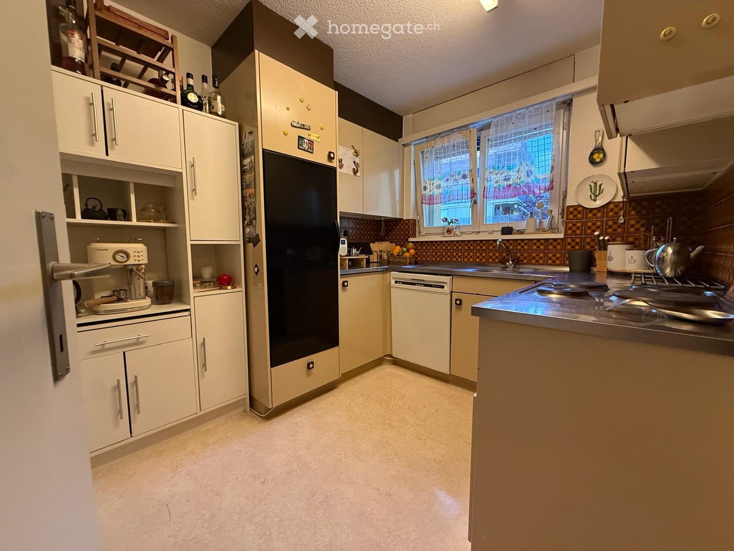 A well-lit kitchen with white cabinets, a black refrigerator, a dishwasher, a coffee maker, and a stove. The countertops are stainless steel, and there is a window with a view of the outside.