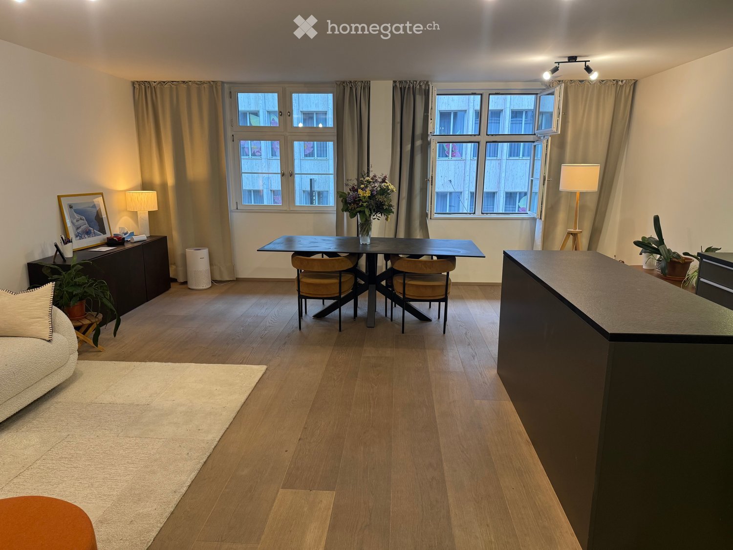 A modern, well-lit living room with wooden flooring, large windows, a sofa, a coffee table, a dining table with chairs, a cabinet, and decorative plants.