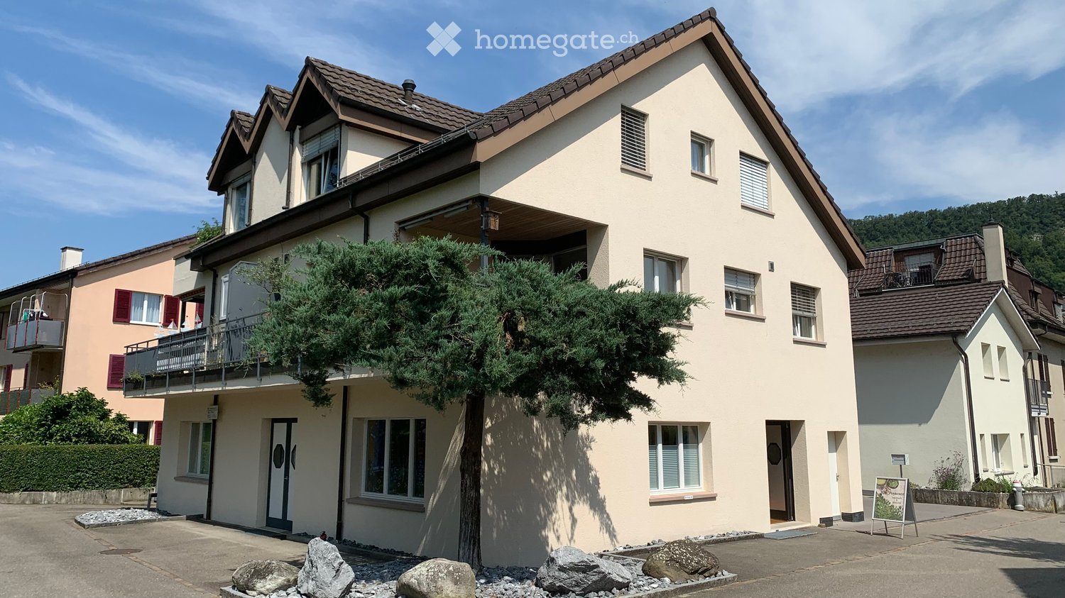 Two-story house with beige walls, multiple windows, and a balcony on the upper floor. The house features a well-maintained garden with a large evergreen tree and decorative rocks in the foreground.