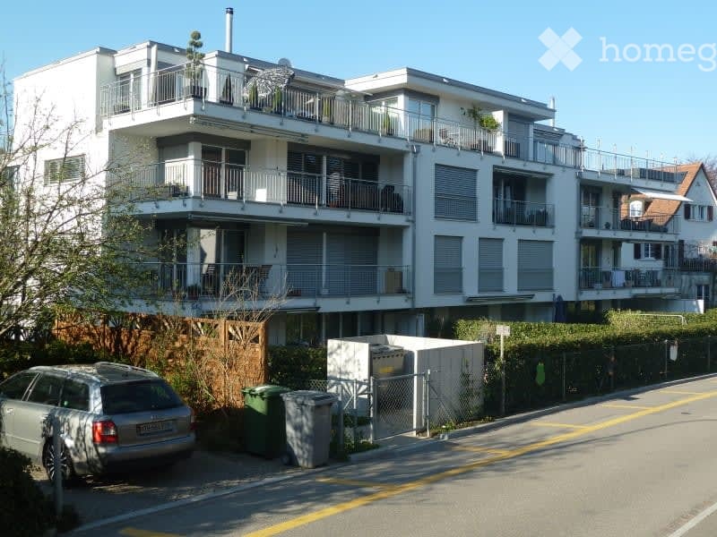 Modern multi-story apartment building with balconies, white exterior, and surrounding greenery.