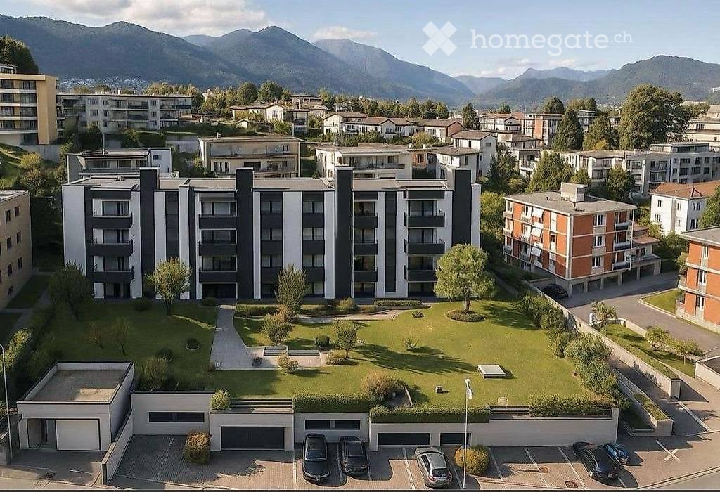 Aerial view of a modern residential complex with multiple apartment buildings, a central green courtyard, and a parking area with several cars.