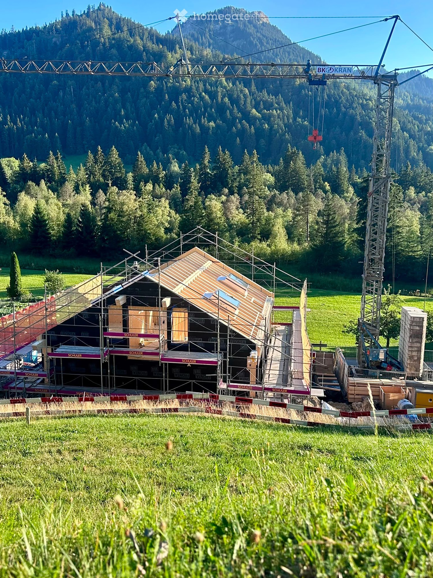 A partially constructed building with scaffolding, surrounded by a grassy field and forested mountains in the background. A crane is visible on the right side.
