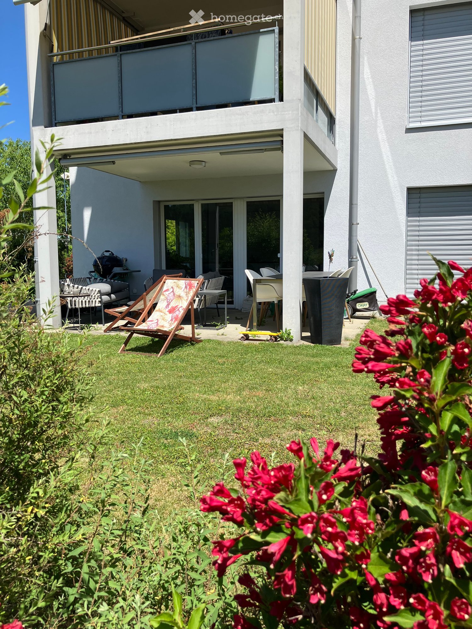 Garden area with a lounge chair, dining table, chairs, and a skateboard near a modern building with balconies.