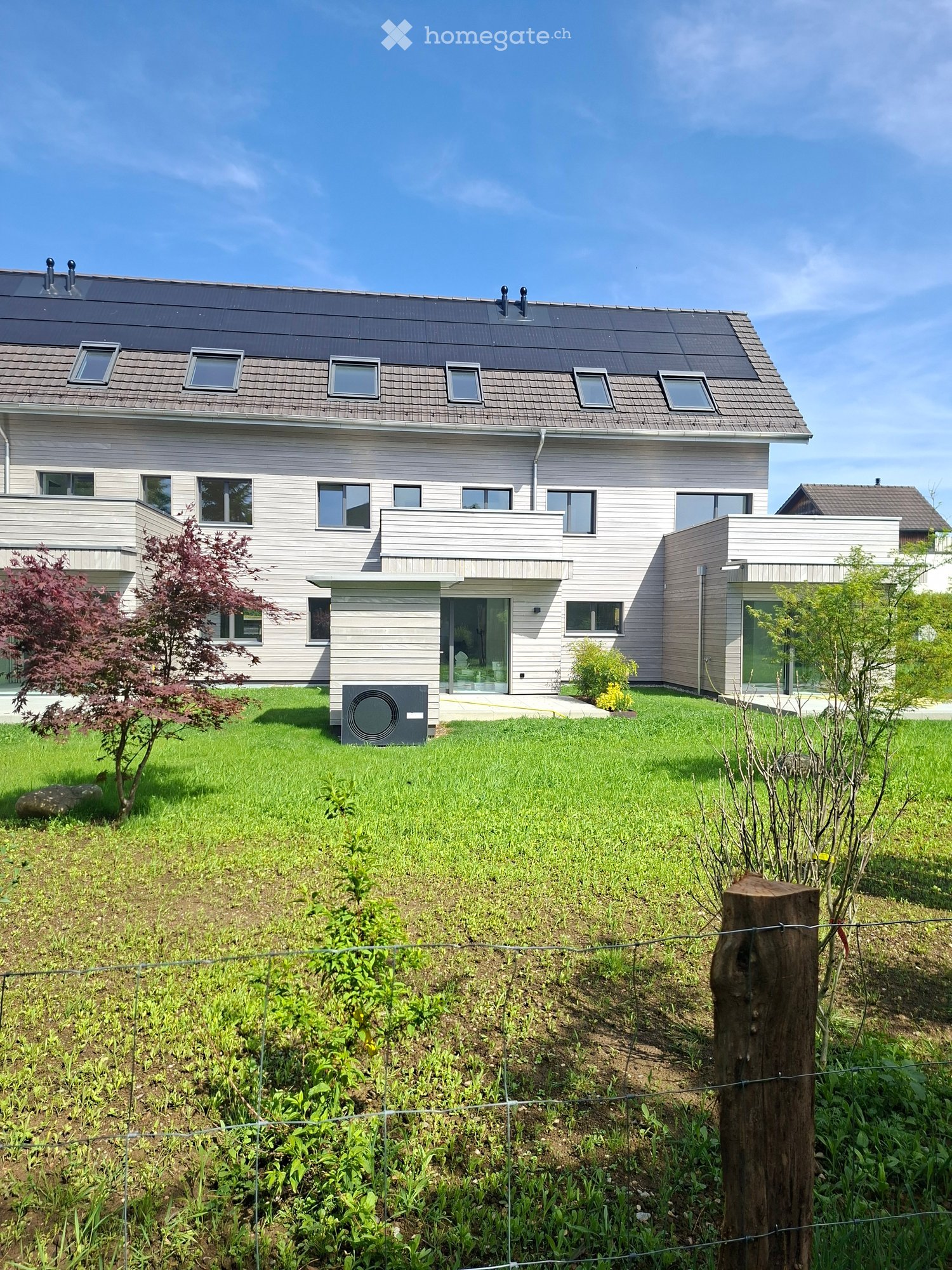 Modern two-story house with grey siding, multiple windows, and a flat roof with skylights. The house features a raised ground floor and is surrounded by a well-maintained lawn and garden area.