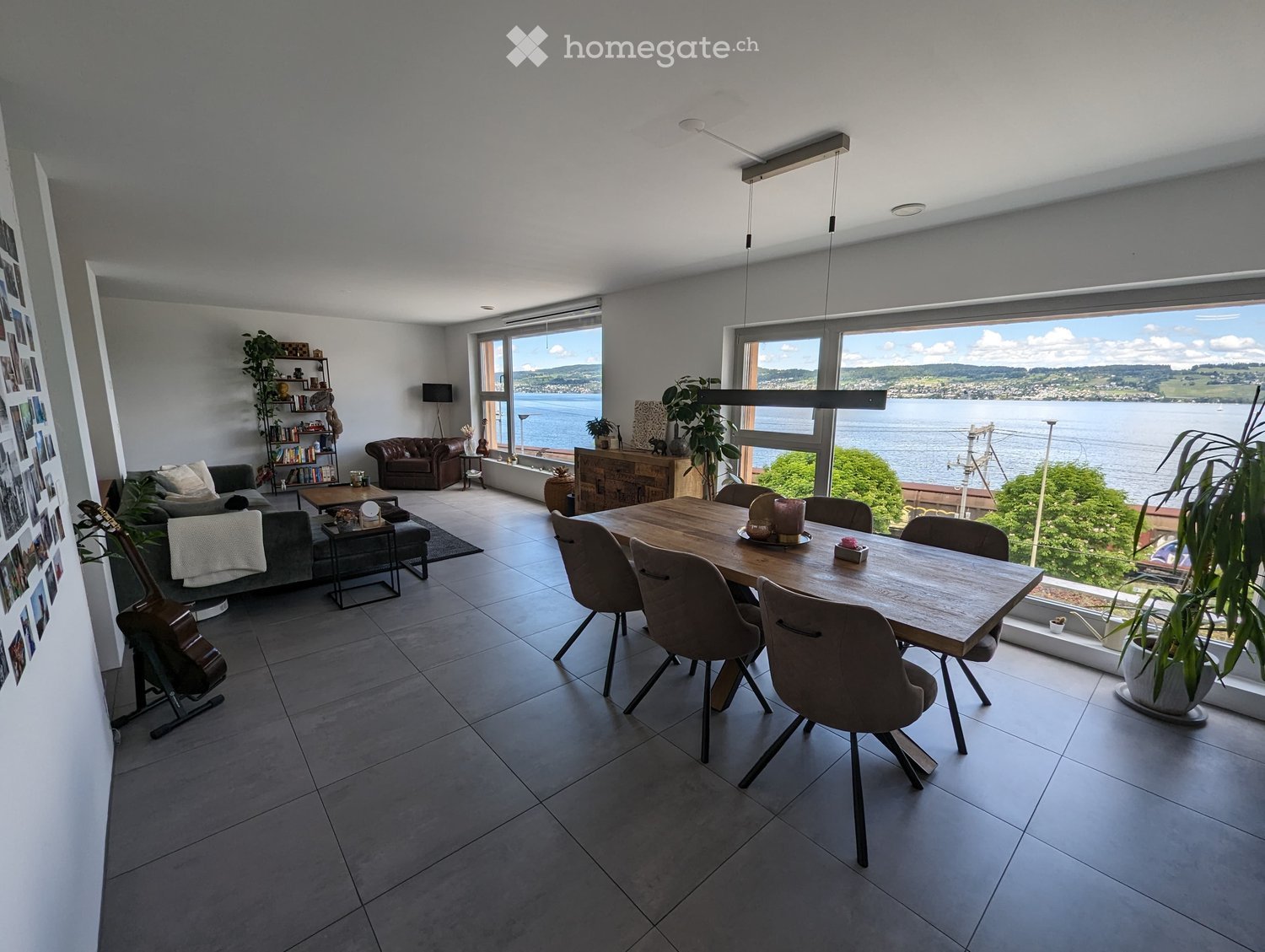Open-plan living and dining area with large windows overlooking a scenic lake view. Features a grey sofa, wooden dining table with six chairs, a guitar, a bookshelf, and several plants.