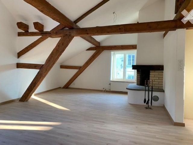 Empty room with exposed wooden beams, wooden floor, white walls, fireplace, and a window letting in natural light.
