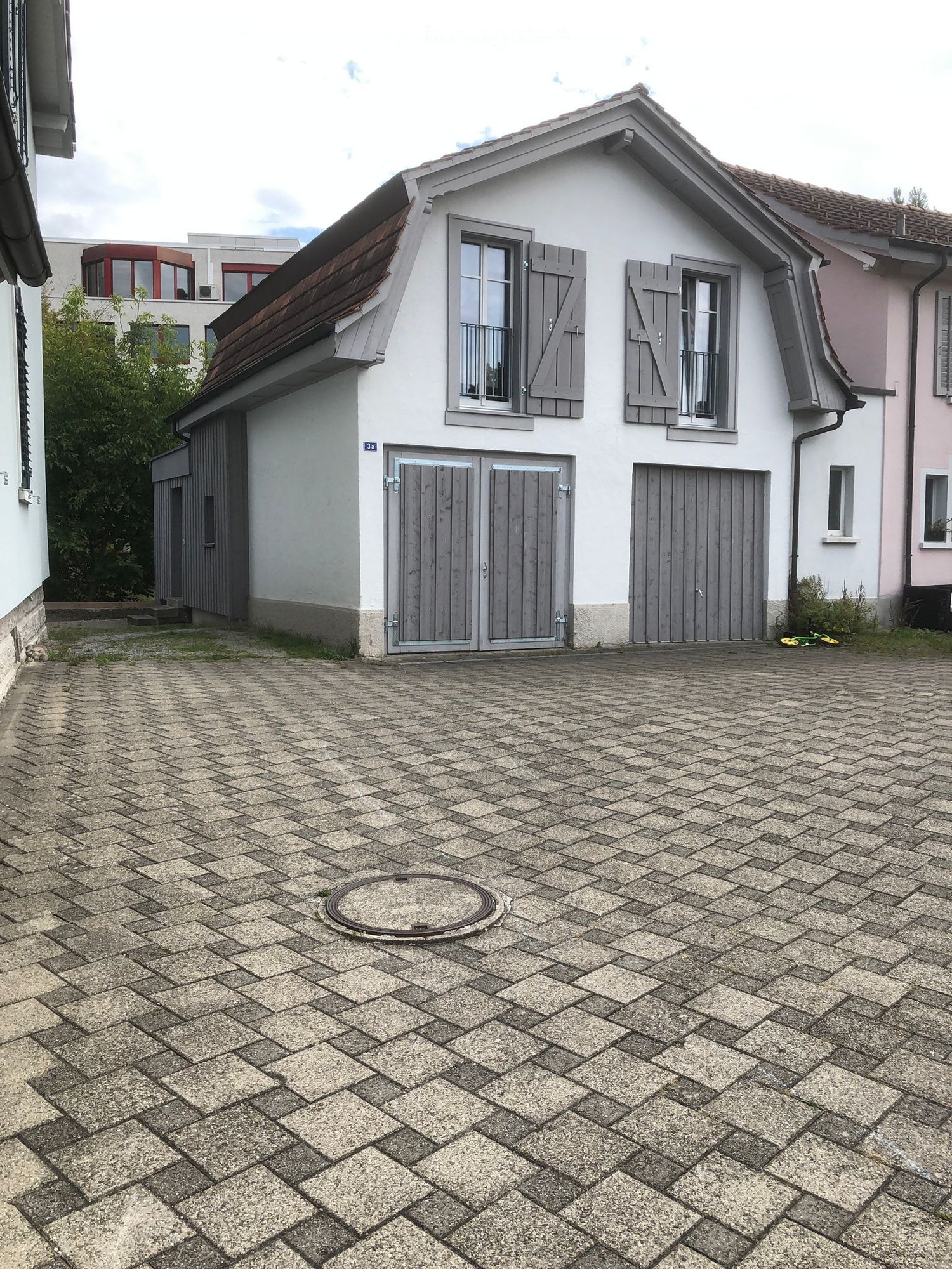 Two-story house with white walls, grey shutters, and a tiled roof. The entrance features double doors, and the driveway is paved with interlocking stone tiles.