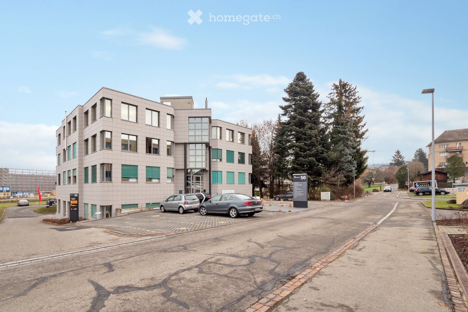 Office building with multiple floors, glass windows, parking lot in front, street with cars parked, trees, street light