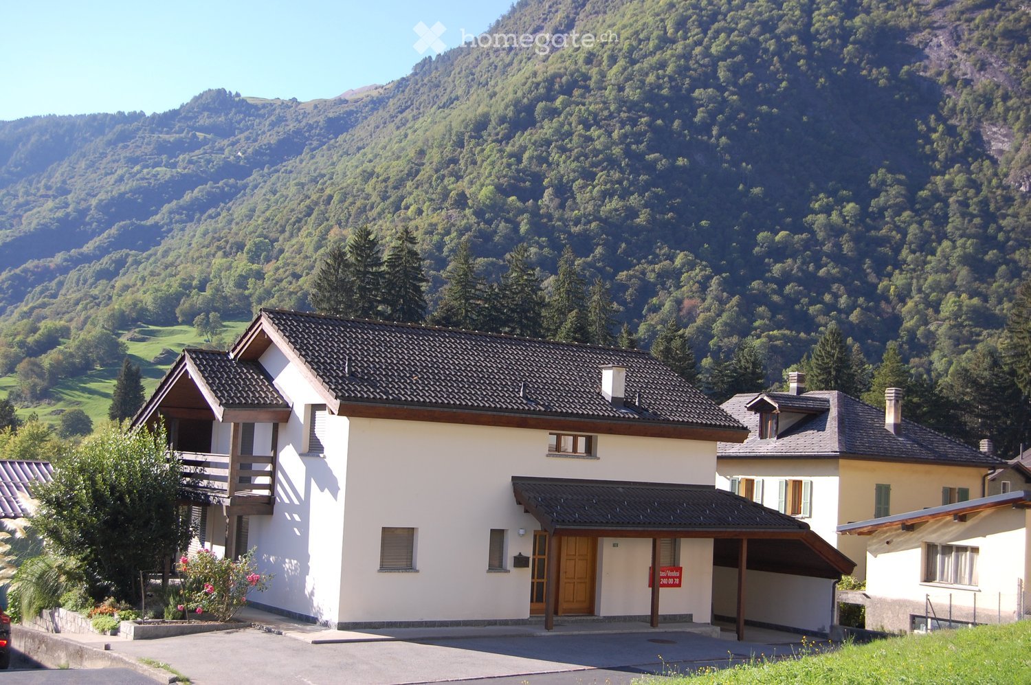 A two-story house with a dark tiled roof, white walls, and a wooden balcony on the left side. The house has multiple windows and a covered entrance with a wooden door. The surrounding area includes a driveway, a small garden with flowers, and a scenic mou